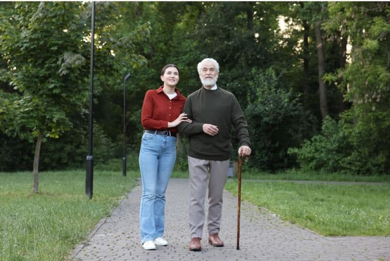 Caregiver walking arm-in-arm with an elderly man using a cane along a park path — caregiver support and self-care